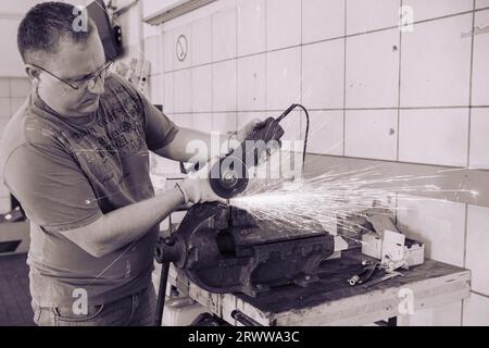 A man works as a grinder in a workshop. A master in glasses grinds a part clamped in a vice with a grinder. Sparks from the metal fly around like a br Stock Photo