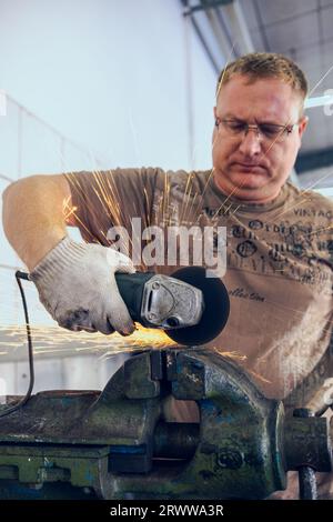 A man works as a grinder in a workshop. A master in glasses grinds a part clamped in a vice with a grinder. Sparks from the metal fly around like a br Stock Photo