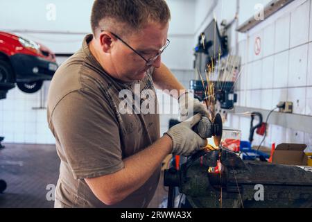 A man works as a grinder in a workshop. A master in glasses grinds a part clamped in a vice with a grinder. Sparks from the metal fly around like a br Stock Photo