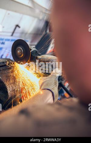 A man works as a grinder in a workshop. A master in glasses grinds a part clamped in a vice with a grinder. Sparks from the metal fly around like a br Stock Photo