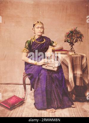 A formal studio portrait of an Indian woman from Travancore. She is dressed in a sari with ornate gold jewellery, indicating she is from a high caste, and holds a handbag. She sits on a chair with one arm resting on top of a book on a side table with a flower arrangement. There is another book to her right on the floor. The photograph has been hand-painted with bright colours. Handwritten caption reads: A Travancore Lady. Stock Photo