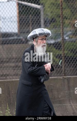 An older Jewish man walks during a light drizzle wearing a homemade rain hat. On Lee Avenue in Williamsburg, Brooklyn, New York. Stock Photo