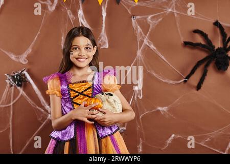 cheerful girl in Halloween costume holding pumpkins and skull on brown backdrop, spooky season Stock Photo