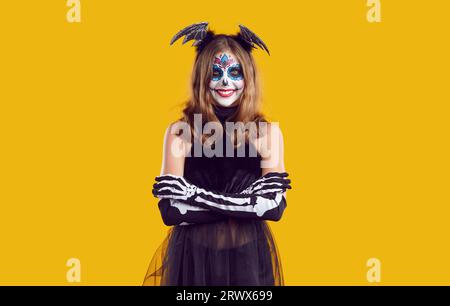 Smiling girl in creative skeleton costume and bat wings on her head with skull makeup, crossed her arms over her chest. Stock Photo