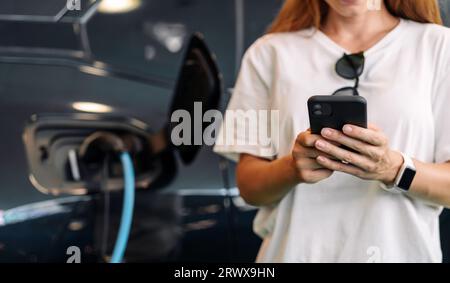 Female person using her mobile phone for charging her electric car. EV station and mobile paying concept. Stock Photo