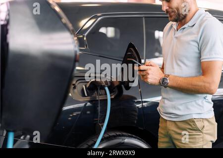 Young adult man using  mobile paying for charging EV. Businessman wearing casual clothing using his cellphone in front of black electric car. Stock Photo