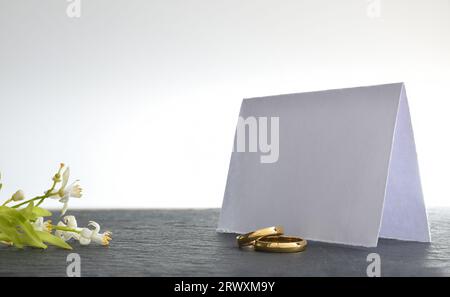 Two engagement rings on slate table with blank paper card behind and flower bouquet and white isolated background. Front view. Stock Photo