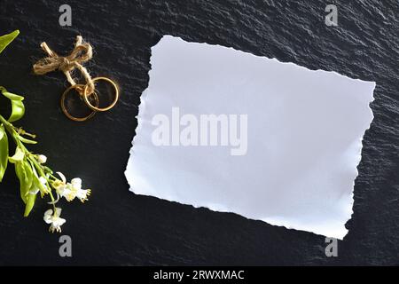 Paper white card on black slate background with rings bound with jute rope and flowers. Top view. Stock Photo