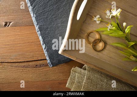 Two engagement rings on a wooden tray with flowers on rustic wooden table decorated with slate stone and burlap cloth. Top view. Stock Photo