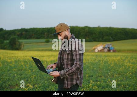 Caucasian young male farmer in hat standing in field and typing on keyboard of laptop computer. Attractive man using device in margin in summer and br Stock Photo