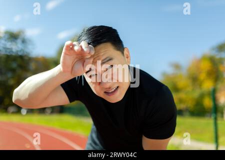 Close-up photo. Portrait of a young Asian male sportsman tiredly bent over at the stadium, resting after jogging and training, looking at the camera. Stock Photo