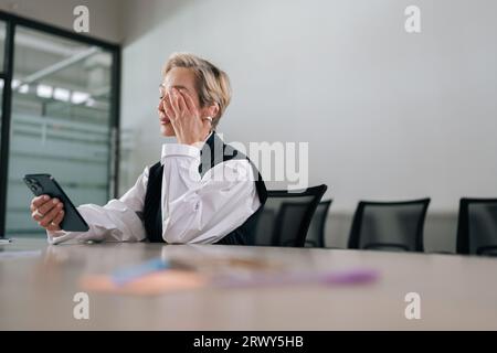 Low-angle view of tired middle-aged businesswoman using mobile phone at office desk, feeling discomfort from dry irritated eyes. Stock Photo