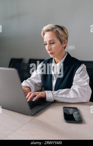 Vertical shot of confident middle-aged 50s businesswoman in suit sitting at modern office desk working on laptop do freelance online job. Stock Photo