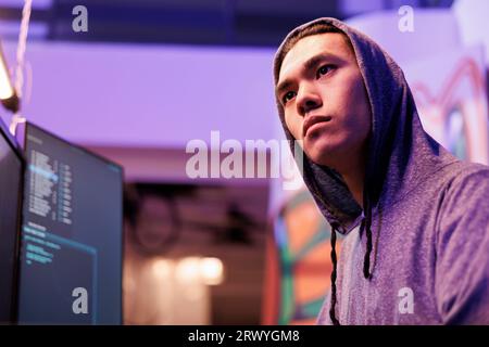 Concentrated asian criminal coding malware on computer while breaking law. Young focused hacker in hood hacking database server and using malicious software to steal data Stock Photo