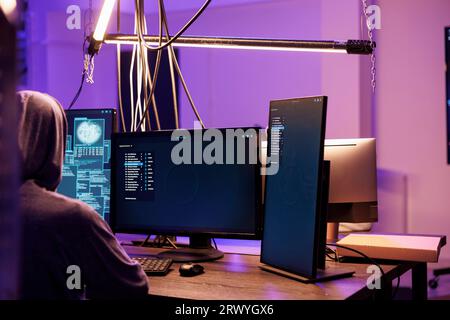 Hacker running malicious software on screen for cracking password. Malware code displaying on multiple monitors with criminal in hood working on computer is abandoned warehouse Stock Photo