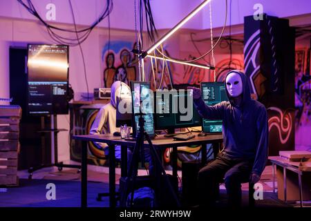 Anonymous scammer live streaming threat on mobile phone while breaking law in abandoned warehouse. Hacker in hood recording ransom video for victim on smartphone late at night Stock Photo