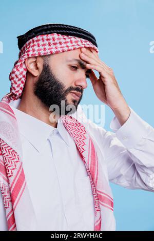 Arab man in ghutra headscarf holding hand on forehead while suffering from headache. Muslim person in pain dressed in islamic thobe and headdress having high blood pressure symptom Stock Photo