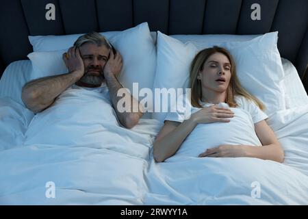 Irritated man with headphones lying near his snoring wife in bed at home Stock Photo