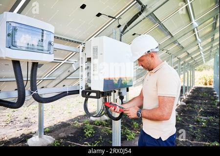 Good worker checking solar panel voltage. Turning on solar system. Man in helmet with current clamps in hands on back side of battery. Stock Photo