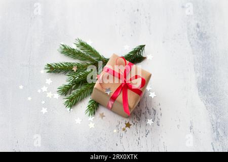 A small gift wrapped in kraft paper with a red ribbon on a light blue background with natural Christmas tree branches, surrounded by a shiny confetti Stock Photo