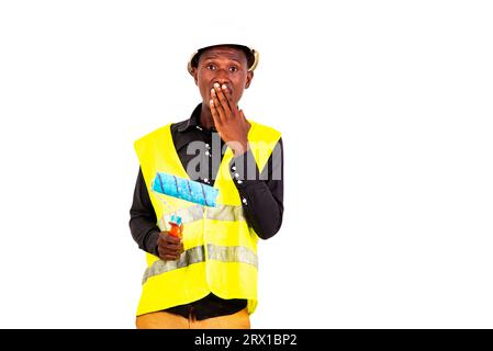 young amazed painter man with hand over mouth wearing green vest and safety helmet holding brush roller. Stock Photo