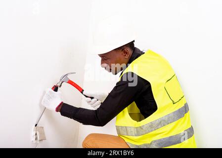 young male electrician wearing white glove and using hammer and screwdriver to install electrical outlets and switch. Stock Photo