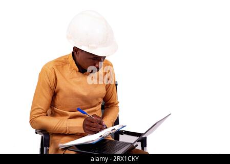 young architect business man wearing white hard hat sitting on chair and writing on clipboard using laptop. Stock Photo