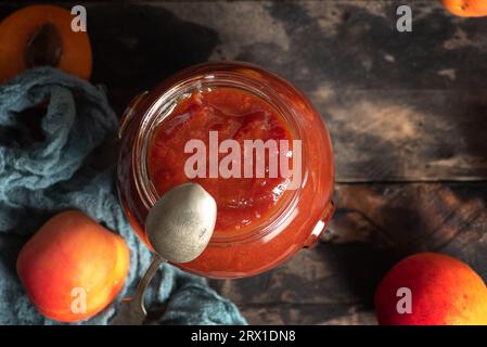 Delicious, homemade apricot jam in a glass jar on a wooden table. There is also a small metal spoon, whole apricot fruits, half fruit. View from above Stock Photo