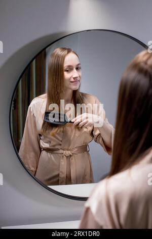 Pretty Woman Combing Her Beautiful Hair With Brush While Standing Near Mirror In Bathroom, Attractive Young Lady Looking To Her Reflection And Smiling Stock Photo