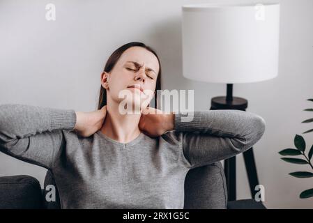 Close up young unhappy caucasian woman sitting on couch holding hands on neck, suffering from neck pain, incorrect posture, joint inflammation. Unheal Stock Photo
