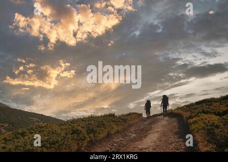 Two hiker silhouettes on the hiking trail high in the mountains. Two men with backpacks and walking poles go towards the horizon in the tcenic sunset Stock Photo