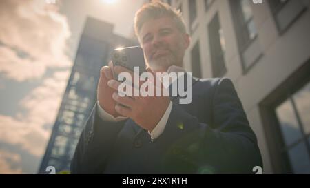 Smiling old man middle aged businessman using mobile phone outdoors in sunshine. Happy senior adult business male user employer holding cellphone Stock Photo