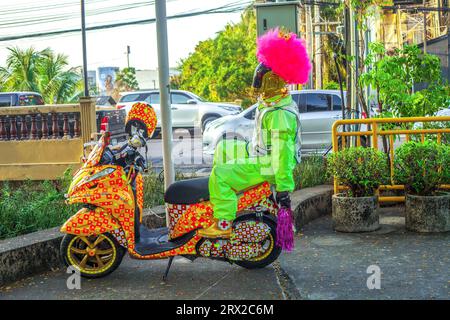 Phuket, Thailand - 03 March 2018: Robot in pink wig, golden crown and green suit sitting on painted scooter on city street. Funny weird mannequin sits Stock Photo