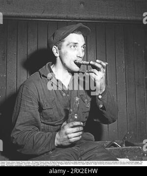 In the 1940s. It is lunch break and a man is eating a sandwich and has a bottle of a drink in his hand. A craftsman who, judging by the appearance of his hands, has a rather dirty job. Period-typical cap with the correct "twist" and with a wooden tool box next to him where he sits. Sweden 1946. Kristoffersson ref X92-4 Stock Photo