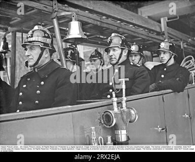 In the 1940s Firemen from the Kungsholmen fire station in Stockholm rush out in their open fire truck. Dressed in fire uniforms and helmets with emblems, they drive out of the fire station. The fire engine is equipped with ladders and a pump equipment. A brass bell sounds the alarm during the call. February 1940. Kristoffersson ref 57-3 Stock Photo