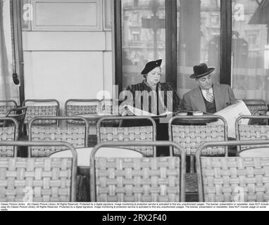 Couple in the 1950s. A couple pictured sitting in chair on an outdorr café, with empty seats except the two. The are reading the daily paper. She is dressed in a fur coat and matching hat, he in a suit with a coat over and a hat. Sweden 1957 Stock Photo