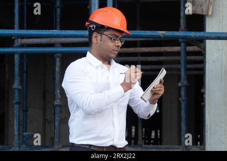 Male engineer in white shirt and hardhat writing notes at construction site. He is focused holding notepad and pen Stock Photo