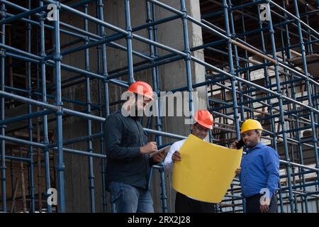 Male architect analyzing blueprint with one of his colleague taking notes and the other one is talking on mobile phone Stock Photo