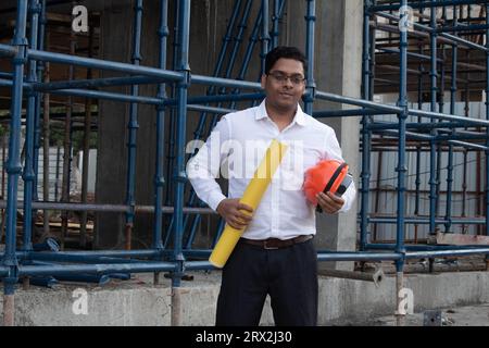 Indian male engineer or architect wearing formalwear holding blueprint and hardhat at construction site Stock Photo