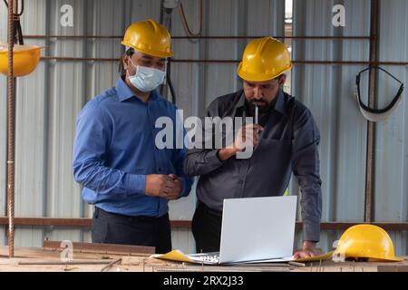 Two construction civil engineers in formalwear and hardhats discussing project under a shelter at site. They are using laptop and blueprint. Stock Photo
