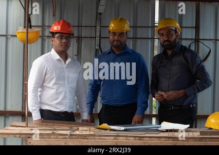 Team of three male architect engineers wearing hardhats standing in a building site. Laptop and blueprint on makeshift table. Stock Photo