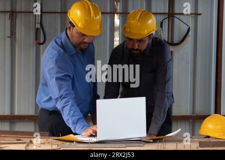 Two male construction architects using laptop and blueprint at project site. They are wearing safety helmet looking at the screen. Stock Photo
