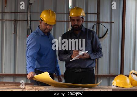Civil engineer holding a blueprint while colleague is taking notes into notepad. They are wearing safety helmet and focused on planning the next phase Stock Photo