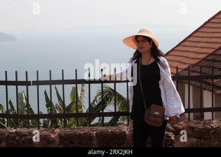 Beautiful young woman in sun hat leaning on fence while standing at terrace. Female traveler spending leisure time outdoors during vacation Stock Photo