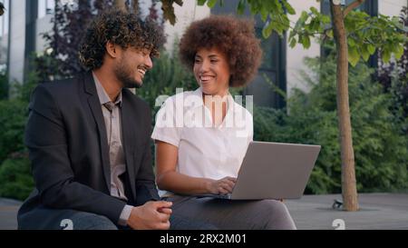 Two diverse multiracial multiethnic business colleagues partners workers employers coworkers sit in city downtown outdoors together with computer Stock Photo