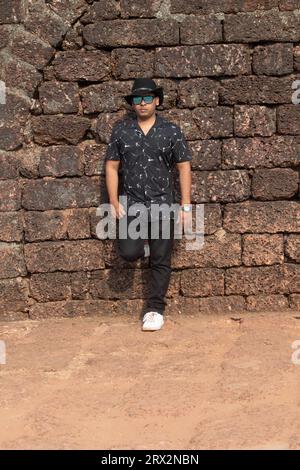 Handsome man in cowboy hat leaning against ancient stone wall. Stylish male traveler in summer hat and sunglasses at historic site Stock Photo