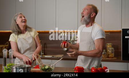 Happy Caucasian senior old couple having fun cooking at home funny man fooling around juggling tomatoes smiling woman cook fresh vegetable salad in Stock Photo