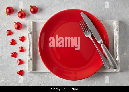 Empty red plate on a wooden tray, red Christmas balls, hearts and cutlery on a gray stone table. New Year's menu Stock Photo