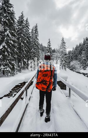 Man tourist and climber with orange backpack standing on the bridge with amazing view on snowy mountains in winter time. Backpacker in snowy weather e Stock Photo