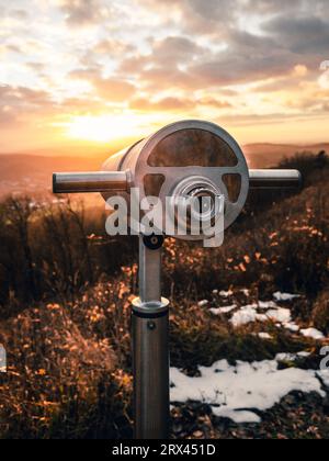 Close up image of a binoculars (telescope) on the hill in nature with beautiful sunset. Photo of outdoor tourist telescope in winter forest with city Stock Photo